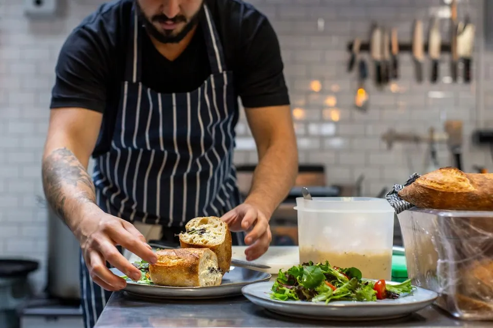 Male chef wearing a striped apron arranges a gourmet baguette sandwich on a plate alongside fresh salad inside a professional restaurant kitchen setting.