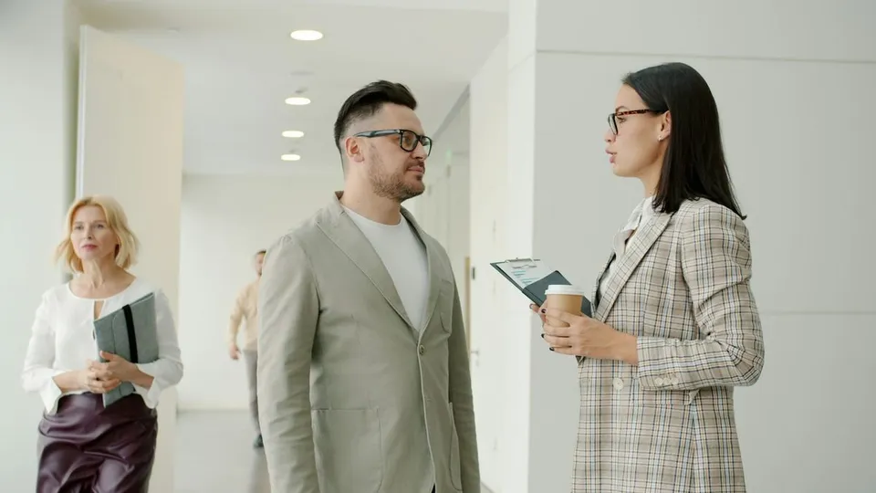 Colleagues in professional attire talk in a bright office hallway, with a woman holding a clipboard and coffee cup while speaking to a man wearing glasses.