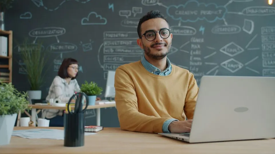 Smiling young man wearing eyeglasses looks forward while working on a laptop at a wooden desk in a modern office with chalk diagrams behind him.