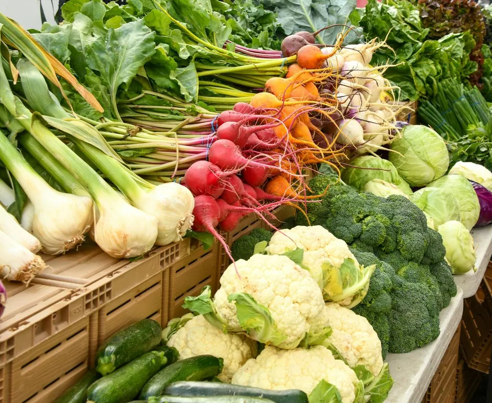 Assorted fresh vegetables including cauliflower, broccoli, radishes, carrots, spring onions, zucchini, and cabbage lie on crates at an open farmer market stand.