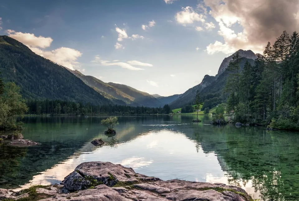 Hintersee lake in Ramsau near Berchtesgaden reflects forested mountain slopes and cloud formations over clear alpine water and rocky shoreline during daytime.