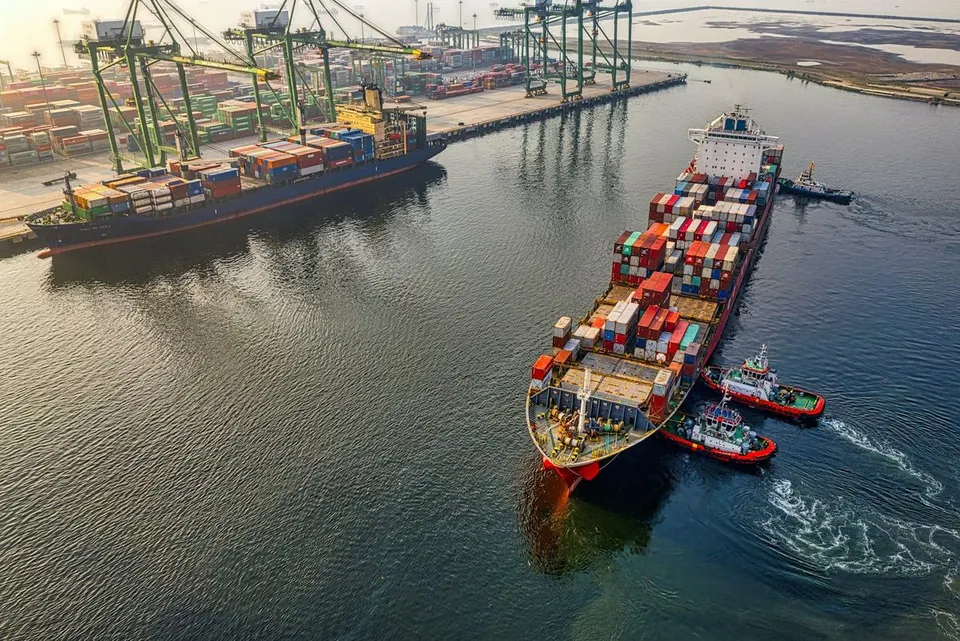 Aerial view shows container cargo ships navigating harbor waters with tugboats assisting, beside busy industrial port docks equipped with tall cranes.