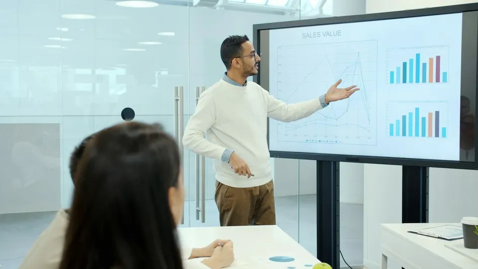 A man wearing glasses explains sales charts displayed on an electronic screen to seated colleagues during an office meeting, pointing toward the graphs with open hands.