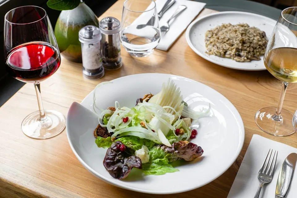 Gourmet salad with sliced pear, walnuts, pomegranate, and lettuce sits in a white bowl alongside risotto, wine glasses, and cutlery on a wooden dining table.