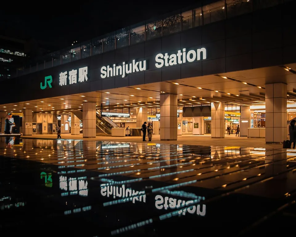 Shinjuku Station facade illuminated at night in Tokyo, Japan, reflects across a dark pool of water while pedestrians walk along the spacious covered modern terminal entrance.