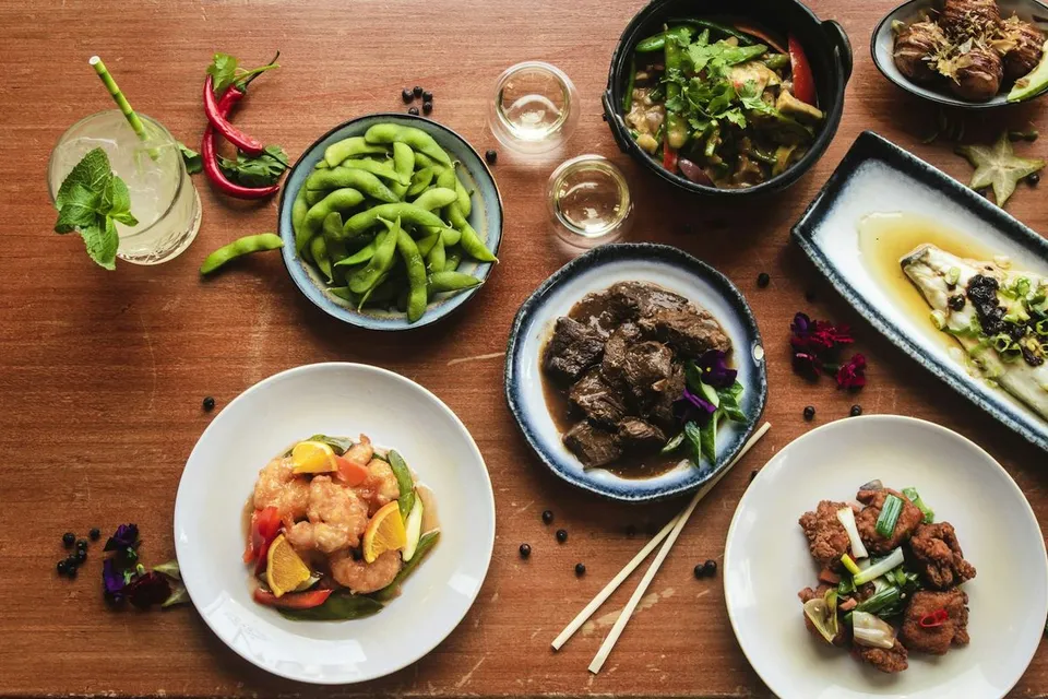 Overhead view of an assortment of cooked Asian dishes, edamame beans, cocktails, and chopsticks arranged neatly across a wooden restaurant dining table, ready for a meal.