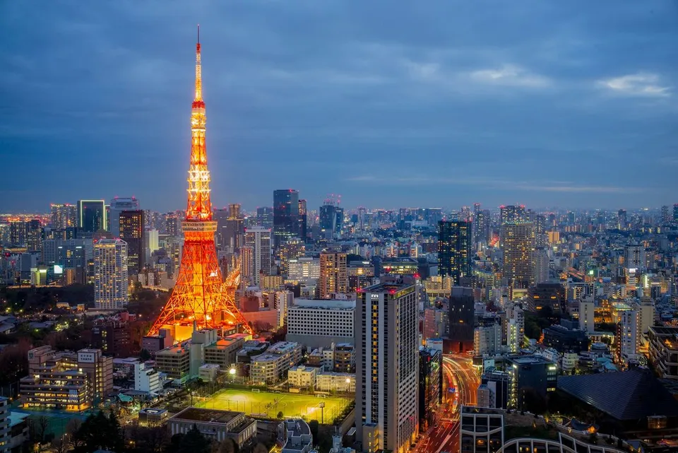 Tokyo Tower shines brightly over the urban cityscape of Tokyo, Japan during twilight, surrounded by modern skyscrapers, street traffic, and an evening sky.