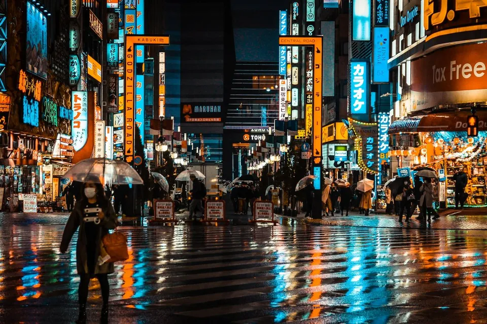 Pedestrians holding umbrellas cross a wet asphalt street reflecting illuminated neon signs along Godzilla Road in Kabukicho, Shinjuku district, Tokyo, Japan.